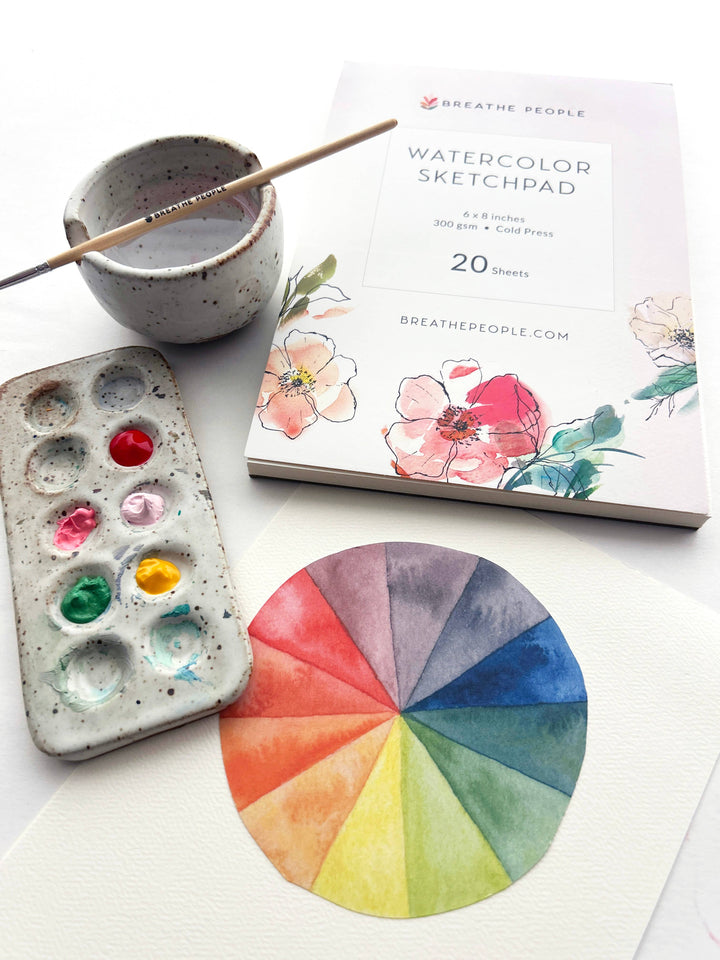 Watercolor sketchpad, color wheel, paint palette, and brush on a white surface