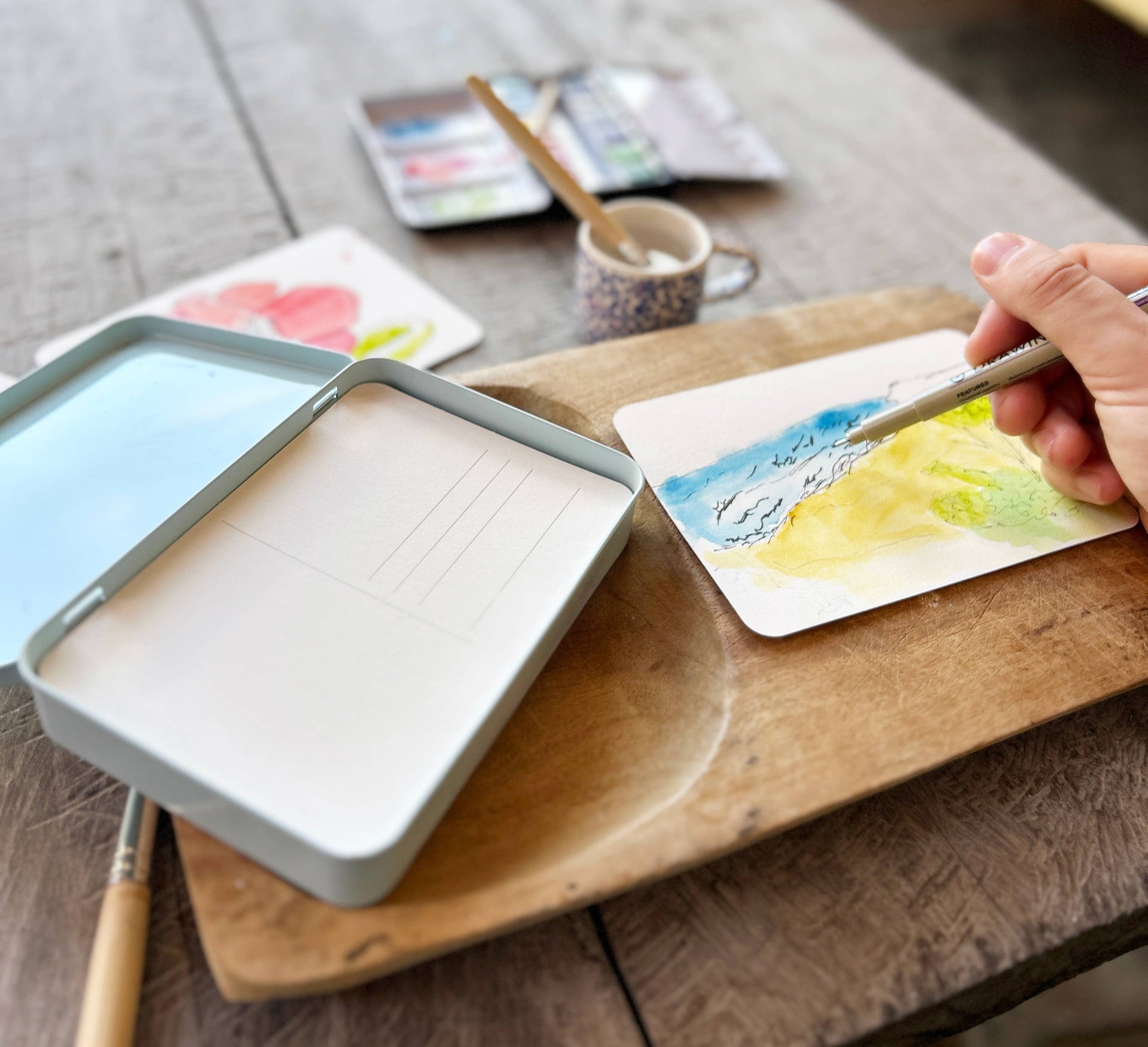 Person painting with watercolors on a wooden table with art supplies.