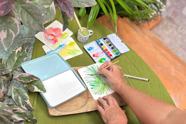 Person painting watercolor flowers on a green table with plants around