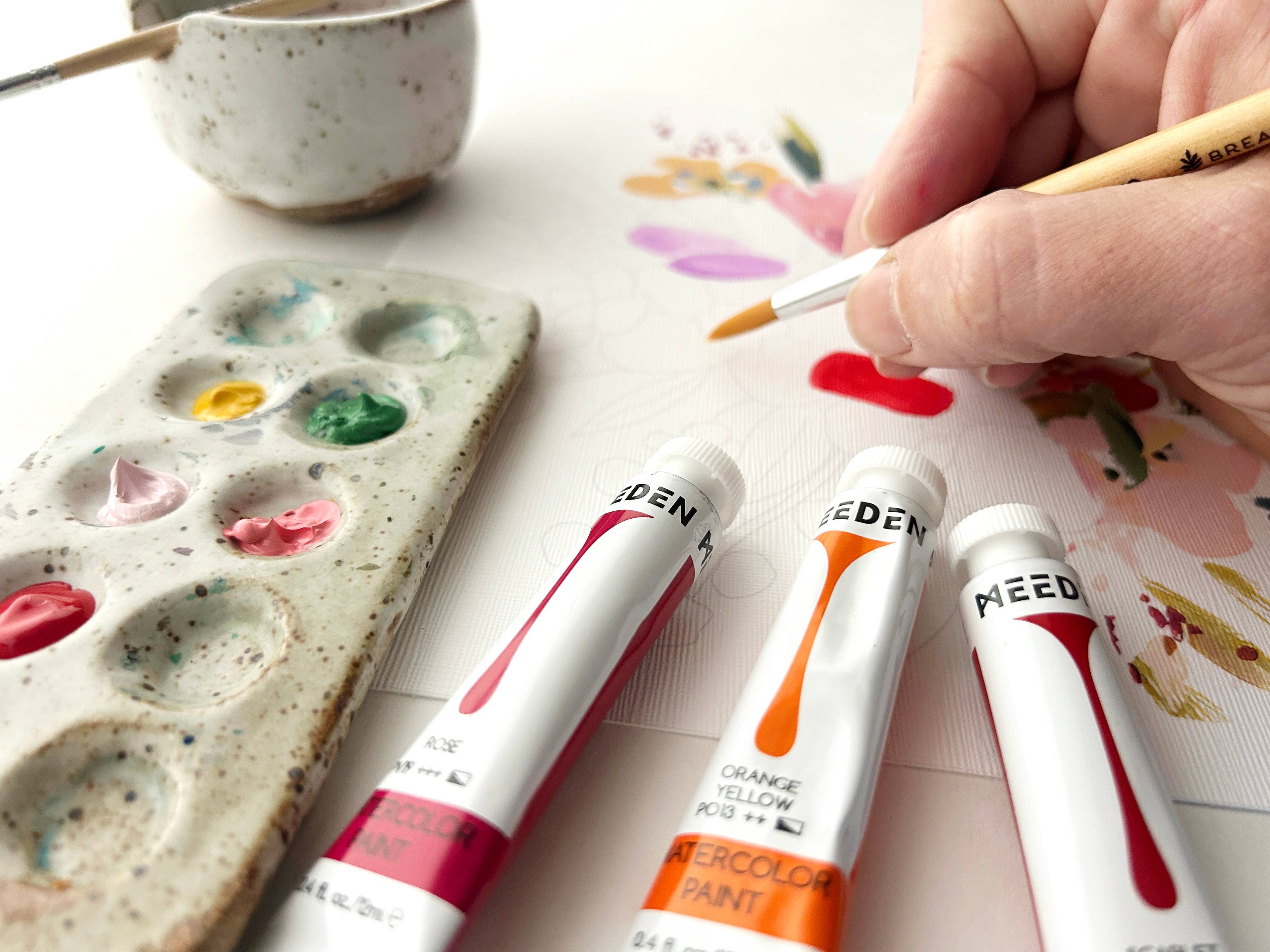 Eden watercolor paint tubes with a hand holding a brush and a paint palette in the background.