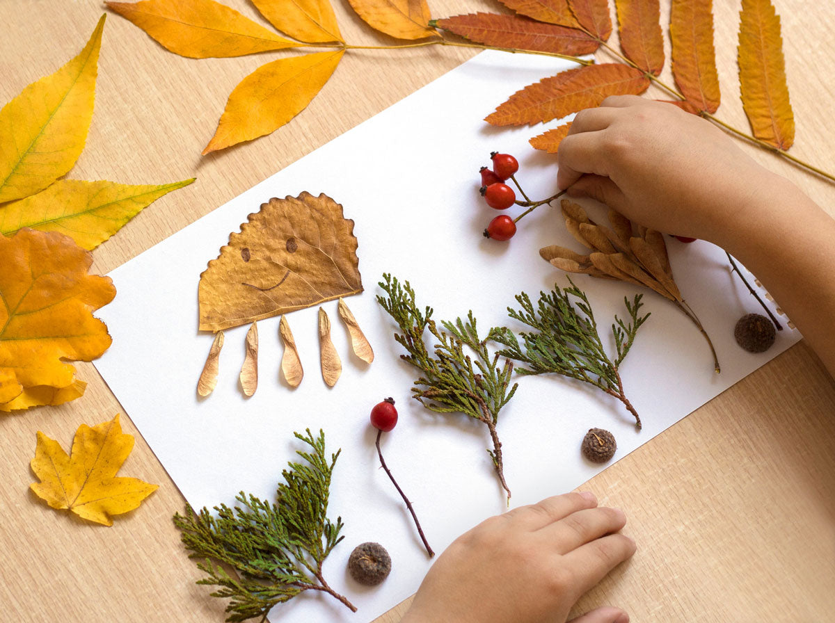 Children's hands arranging autumn leaves and berries on a white sheet of paper.